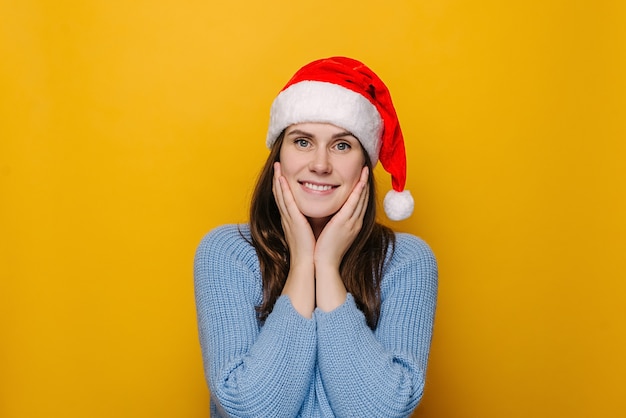 Portrait of happy young woman keeps hands on cheek, cheers coming holidays, looking at camera, dressed in Christmas red hat and winter sweater, isolated over yellow wall. Xmas and good time concept