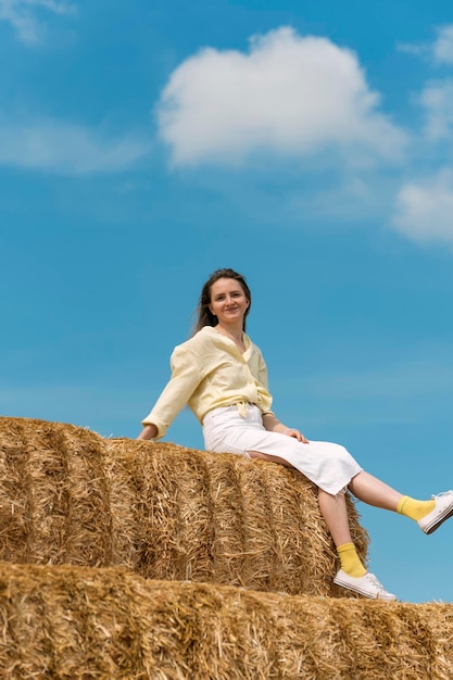 Portrait of happy young woman sitting on high haystack on blue sky background Smiling woman in summer vacation Harvesting