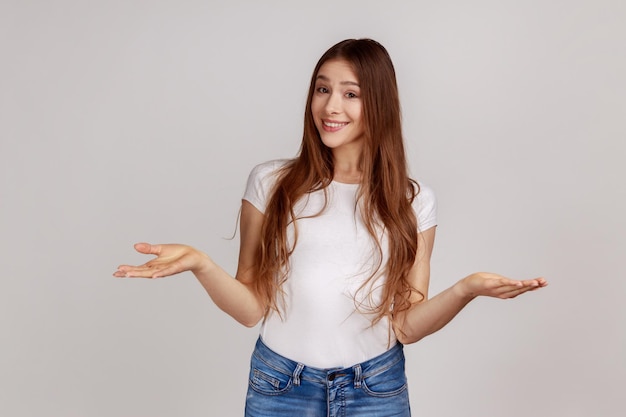 Portrait of happy young woman standing with wide raised arms and looking at camera with toothy smile sharing something, wearing white T-shirt. Indoor studio shot isolated on gray background.