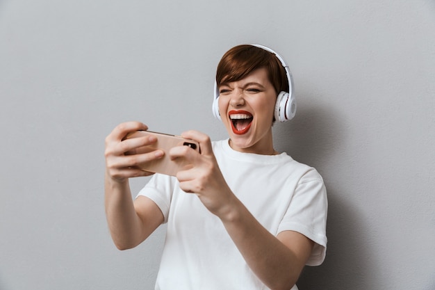 Portrait of happy young woman wearing headphones playing video games on cellphone isolated over gray wall