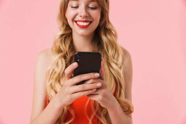 Portrait of happy young woman with long curly hair smiling and holding smartphone isolated over pink wall