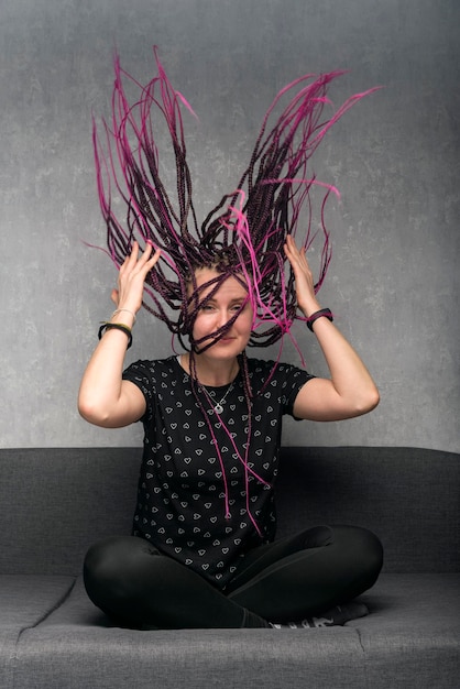 Portrait of happy young woman with long violet dreadlocks sitting on sofa Girl waves her dreads Vertical frame