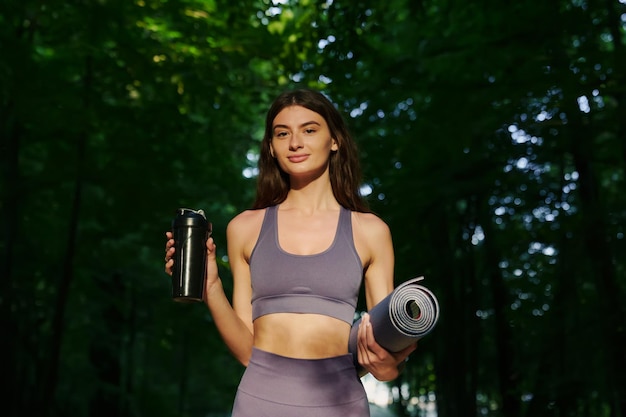 Portrait of a happy young woman with a yoga mat walking through the forest after morning meditation Training outside in summer