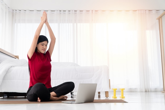 Portrait of healthy young Asian woman practicing yoga exercises sitting in the bedroom and learning online on laptop at home. Concept of exercise and relaxation, Technology for New normal lifestyle