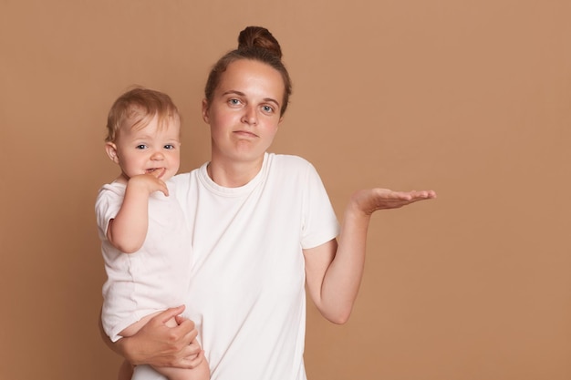 Portrait of helpless confused woman with dark hair wearing white t shirt standing with her baby daughter being not sure spreading hand posing isolated over brown background