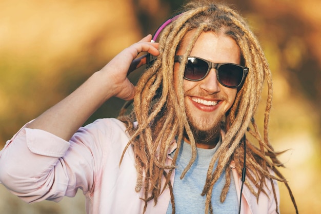 Portrait of hipster blonde bearded boy with dreads wearing headphones and glasses walking smiling and listening his favorite music outdoors an the sunny autumn day