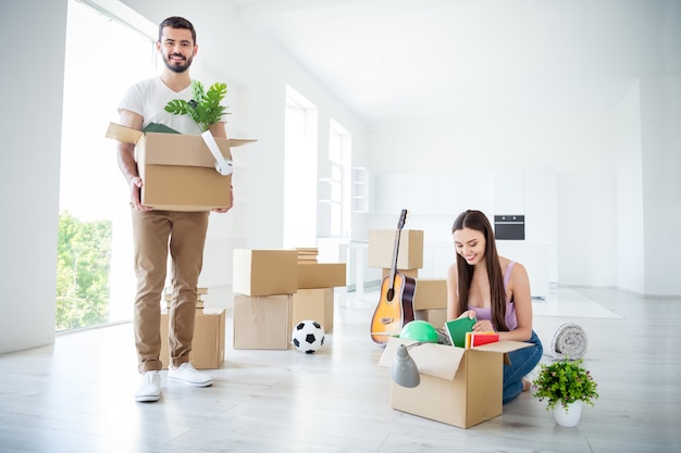 Portrait of his he her she nice attractive lovely cheerful cheery couple packing unpacking stuff unwrapping rental open space flat light white interior house accommodation