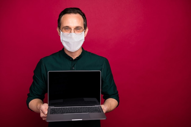 Portrait of his he skilled mature guy holding in hands laptop wearing safety mask mers cov prevention self isolation social distance isolated on bright vivid shine vibrant red color background