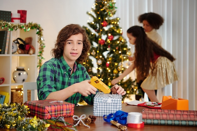 Portrait of Hispanic boy glueing pine cones on wrapped giftbox when his sister decorating Christmas tree