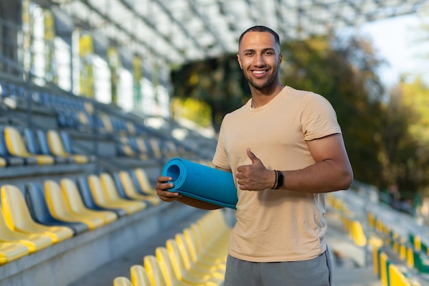 Portrait of hispanic male fitness trainer at sports stadium athlete with yoga mat smiling and