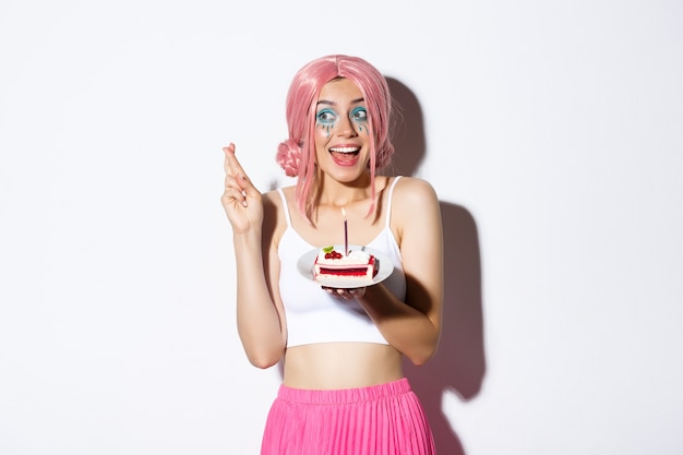 Portrait of hopeful birthday girl in pink wig, making wish with fingers crossed, holding b-day cake, standing over white background