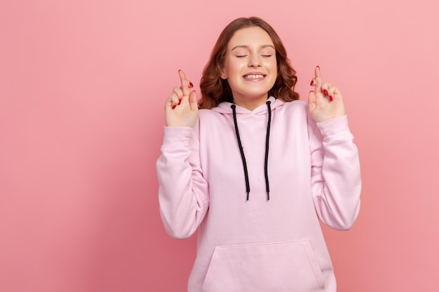 Portrait of hopeful joyous young woman with curly hair in hoodie raising fingers crossed while making wish confident to win Indoor studio shot isolated on pink background