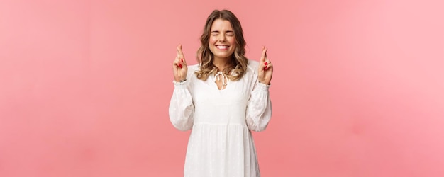 Portrait of hopeful lucky cute blond girl in white dress praying anticiapte miracle making wish to win cross fingers good luck smiling and close eyes and pleading god pink background