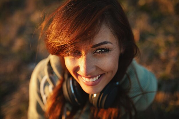 A portrait if a happy smiling woman, with headphones around her neck, is enjoying in sunny day outdoor. Top view.