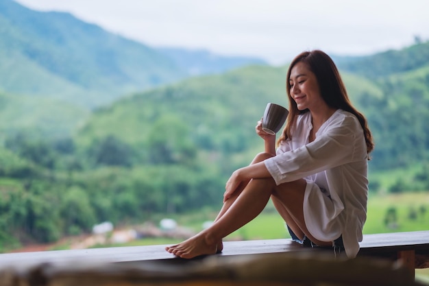 Portrait image of a beautiful asian woman holding and drinking hot coffee on balcony , looking at mountains and green nature