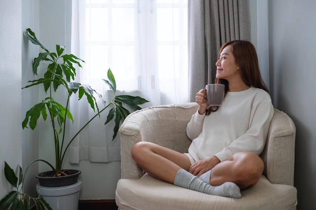 Portrait image of a beautiful asian woman looking outside the window while drinking hot coffee and relaxing on a sofa in bedroom at home