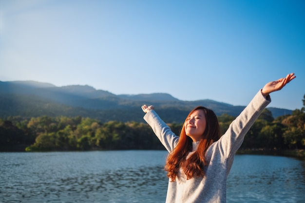 Portrait image of a beautiful asian woman standing and opening arms in front of the lake and mountains on sunny day