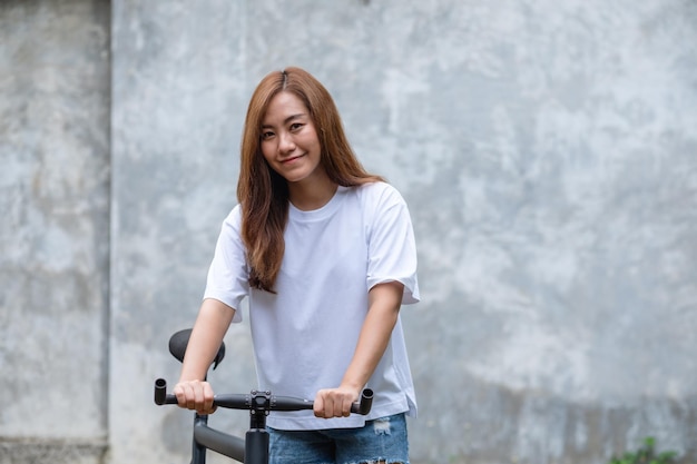 Portrait image of a beautiful young asian woman wearing white tshirt and jean short pant with bicycle and concrete wall background