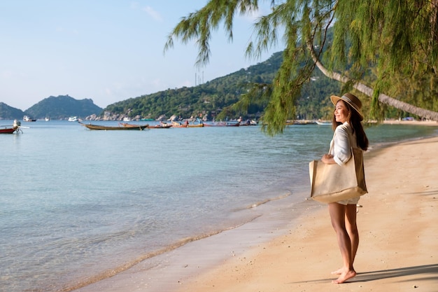 Portrait image of a beautiful young asian woman with hat and bag strolling on the beach