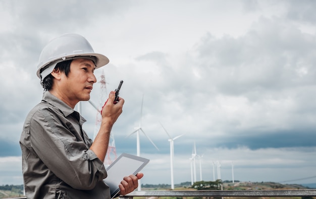 Portrait images of an Asian engineer, technician man standing, holding a tablet and using  radio communication, with Wind turbines, to people and Electricity production concept.