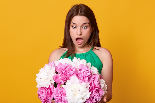 Portrait of impressed worried cute young lady looking at bouquet of flowers with shock, opening mouth and eyes widely