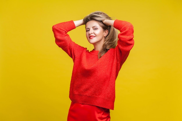 Portrait of impressive happy blonde woman with red lipstick in bright sweater standing with hands in hair looking at camera with seductive smile indoor studio shot isolated on yellow background