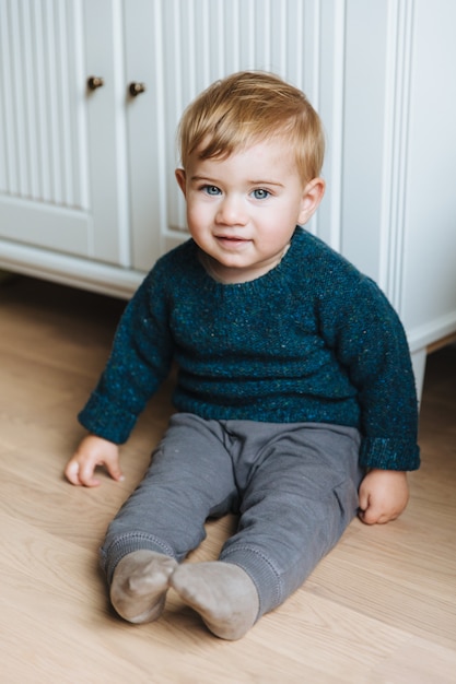 Portrait of infant sits on floor near cupboard, looks with warm blue eyes