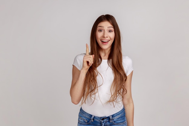 Portrait of inspired woman with long hair pointing finger up with genius idea surprised by suddenly invented smart solution wearing white Tshirt Indoor studio shot isolated on gray background