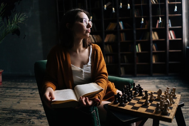 Portrait of intelligent young woman wearing elegant eyeglasses reading book sitting in armchair in dark library room on background of bookcase