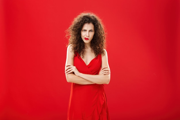 Portrait of intense thoughtful moody woman with curly hairstyle frowning smirking looking from under forehead crossing arms against chest in offended and defensive pose over red background.