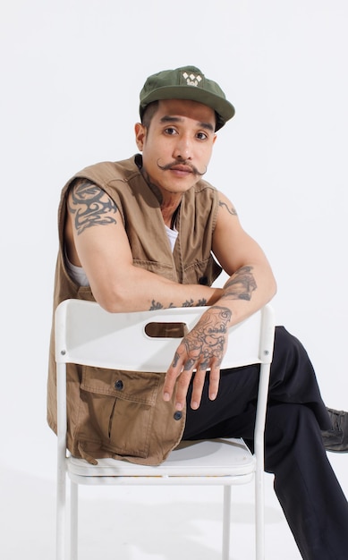 Portrait isolated cutout studio full body shot Asian vintage classy mustache neck arms tattoos male fashion model in casual fashionable safari vest and cap sitting on white chair on white background