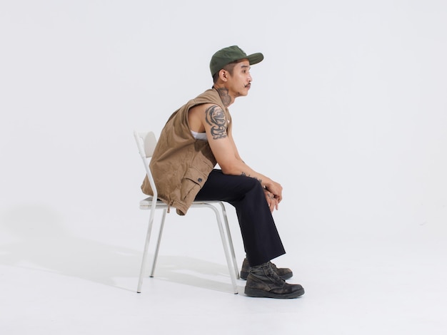 Portrait isolated cutout studio full body shot Asian vintage classy mustache neck arms tattoos male fashion model in casual fashionable safari vest and cap sitting on white chair on white background