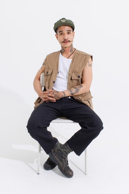 Portrait isolated cutout studio full body shot Asian vintage classy mustache neck arms tattoos male fashion model in casual fashionable safari vest and cap sitting on white chair on white background