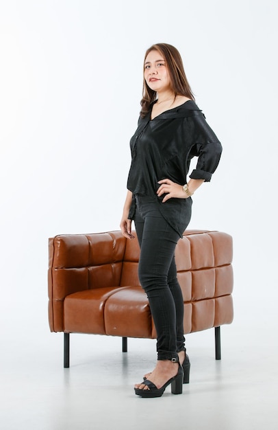 Portrait isolated cutout studio shot Asia cheerful sexy female fashionable model in casual black outfit open shoulder and high heels standing near leather armchair look at camera on white background