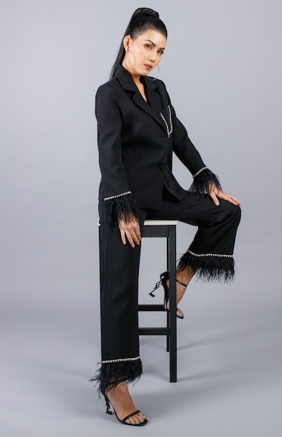 Portrait isolated cutout studio shot of Asian female elegant fashion model in black casual fashionable fur suit and high heels sitting on chair look at camera posing on gray background