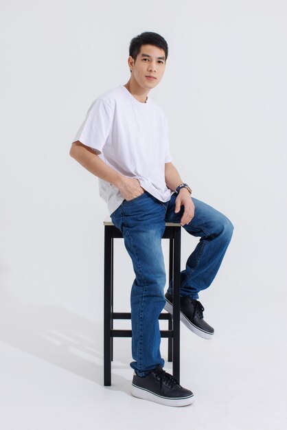 Portrait isolated cutout studio shot Asian handsome cool male fashion model in casual fashionable t shirt and blue jeans sitting on chair holding hand in pocket look at camera on white background