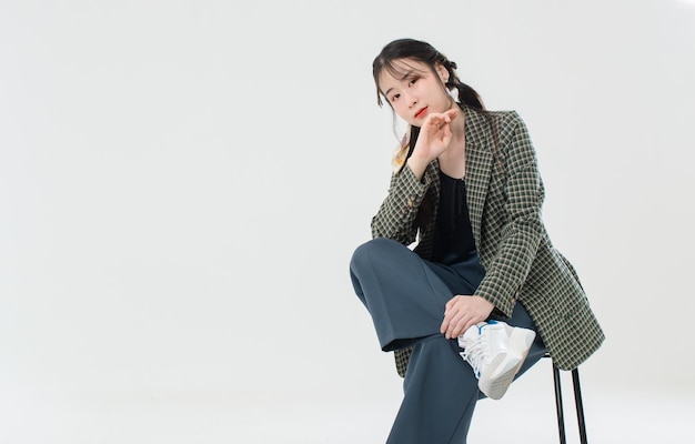 Portrait isolated cutout studio shot of Asian young cute female fashion model with pigtails braids hairstyle in casual plaid suit and crop top shirt sitting on chair posing on white background