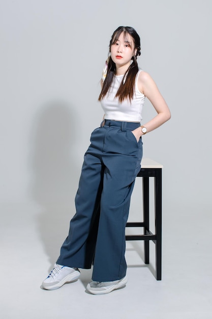 Portrait isolated cutout studio shot Asian young pretty female fashion model with pigtails braids hair in fashionable sleeveless crop top sitting on tall chair posing look camera on white background