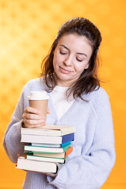 Portrait of jolly woman holding stack of books and coffee cup studio background