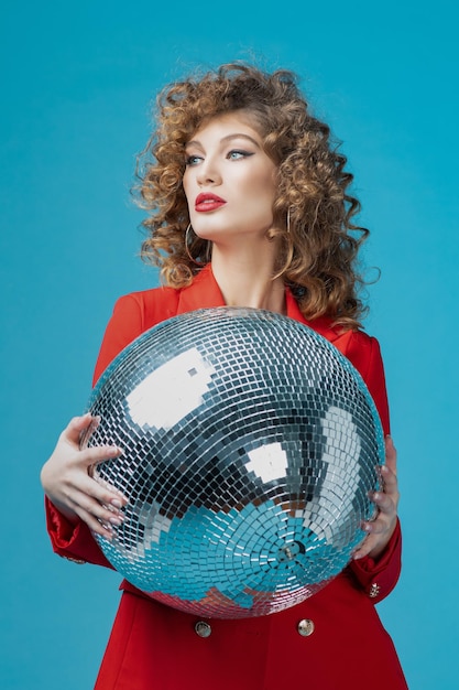 Portrait of a joyful beautiful woman in red dress having a party while standing with disco ball isolated over blue background