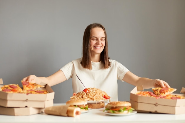 Portrait of joyful charming woman with brown hair wearing white Tshirt sitting at table among different fast food taking pizza from two boxes same time posing isolated over gray background