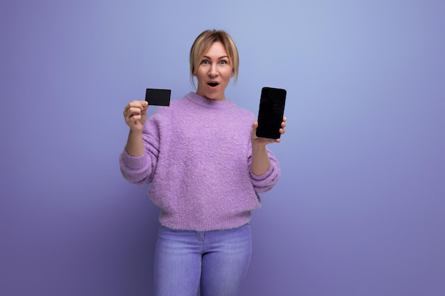 Portrait of joyful cute blondie woman in lavender sweater holding credit card and smartphone with