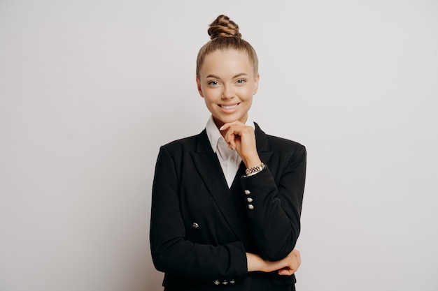 Portrait of joyful female office worker in casual clothes, confident body language with hand on her chin, business woman feeling happy and ready to work, looking forward alone against light wall