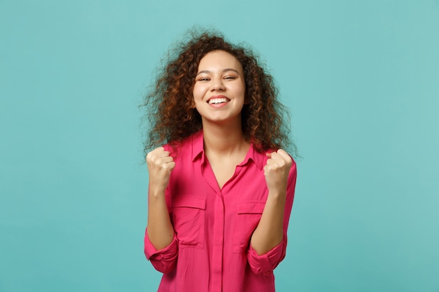 Portrait of joyful funny african girl in pink casual clothes doing winner gesture isolated on blue turquoise wall background in studio. People sincere emotions, lifestyle concept. Mock up copy space.