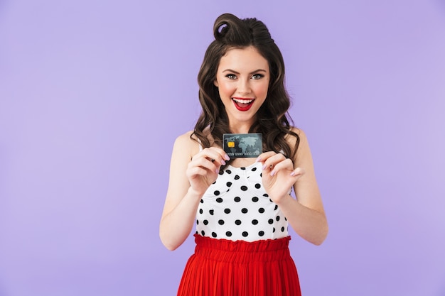 Portrait of joyful pin-up woman in vintage polka dot dress smiling while holding plastic credit card isolated over violet wall