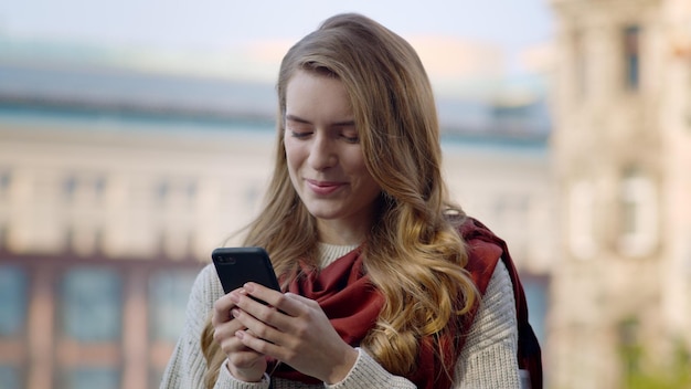 Portrait of joyful woman reading message on mobile phone outdoors Closeup natural beauty girl using smartphone in urban background Smiling female person standing with cellphone on city street