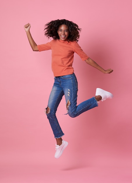 Portrait of a joyful young african woman in orange shirt jumping and celebrating over pink.