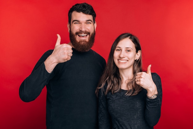 Portrait of joyful young couple standing over red wall and showing thumbs up