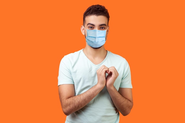 Portrait of joyous handsome brunette man with surgical medical mask in love making heart gesture with hands and smiling broadly at camera indoor studio shot isolated on orange background