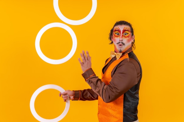 Portrait of a juggler in a vest with a painted face juggling hoops on a yellow background